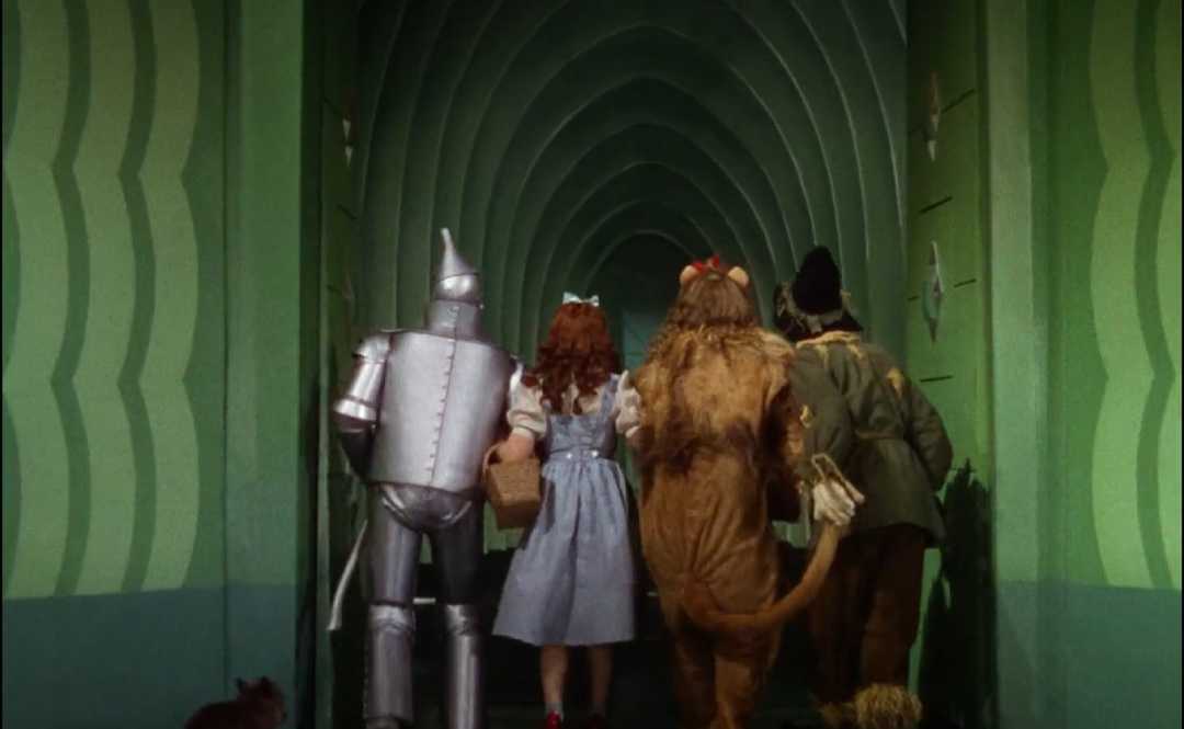 Four characters—the Tin Man, Dorothy, the Cowardly Lion, and the Scarecrow—walk hand in hand down a green hallway, seen from behind, in a scene from "The Wizard of Oz.