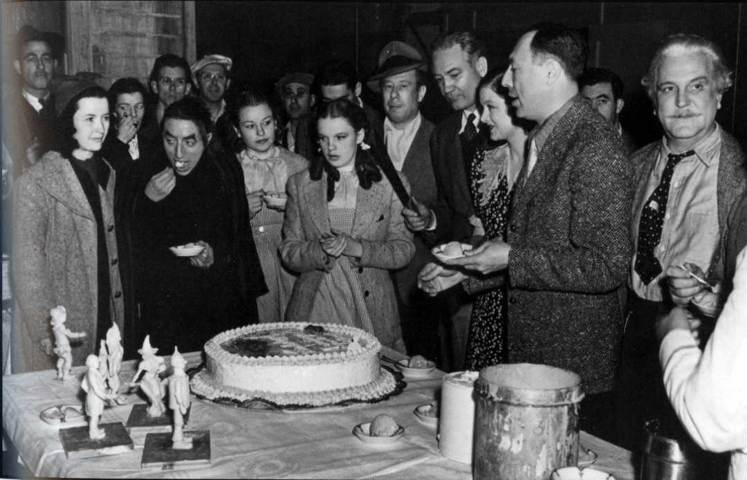 A group of people, some holding plates, gather around a large decorated cake on a table. Several small figurines are arranged in front of the cake, and many of the people appear to be watching or about to eat.