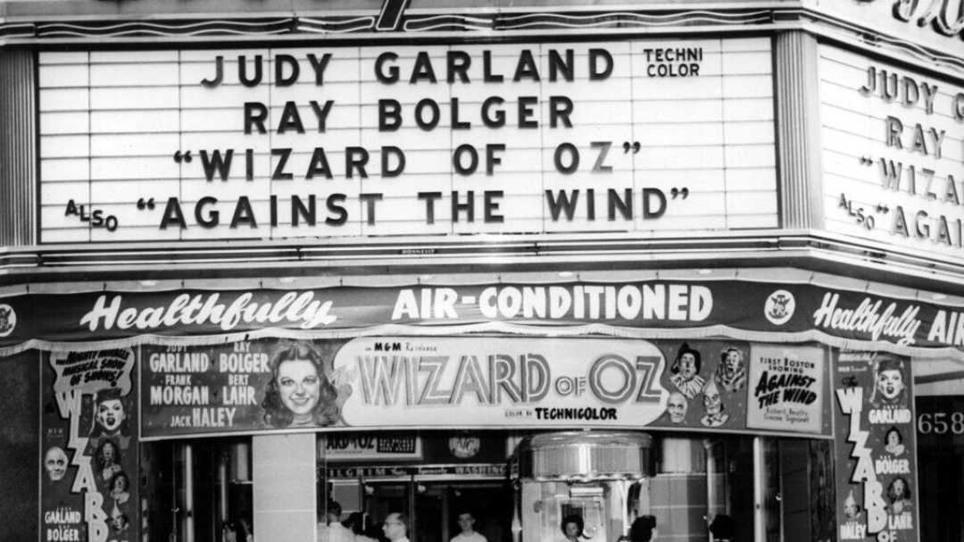 Black-and-white photo of a theater marquee advertising "The Wizard of Oz" starring Judy Garland and Ray Bolger, and "Against the Wind." Posters and signs decorate the entrance, and "air-conditioned" is displayed above the doors.