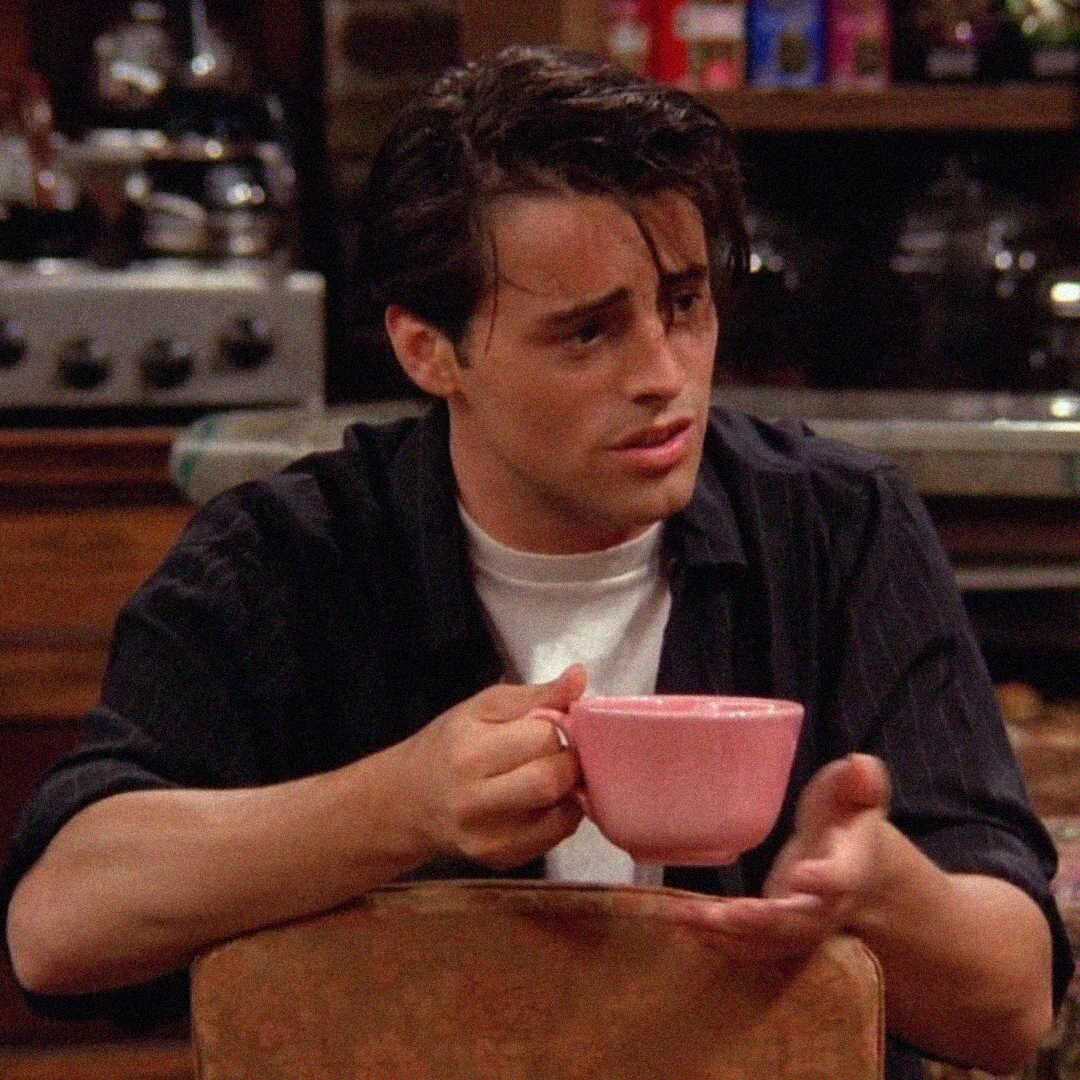 A young man with dark hair holds a pink mug, looking confused or questioning, while sitting in a cozy kitchen setting.
