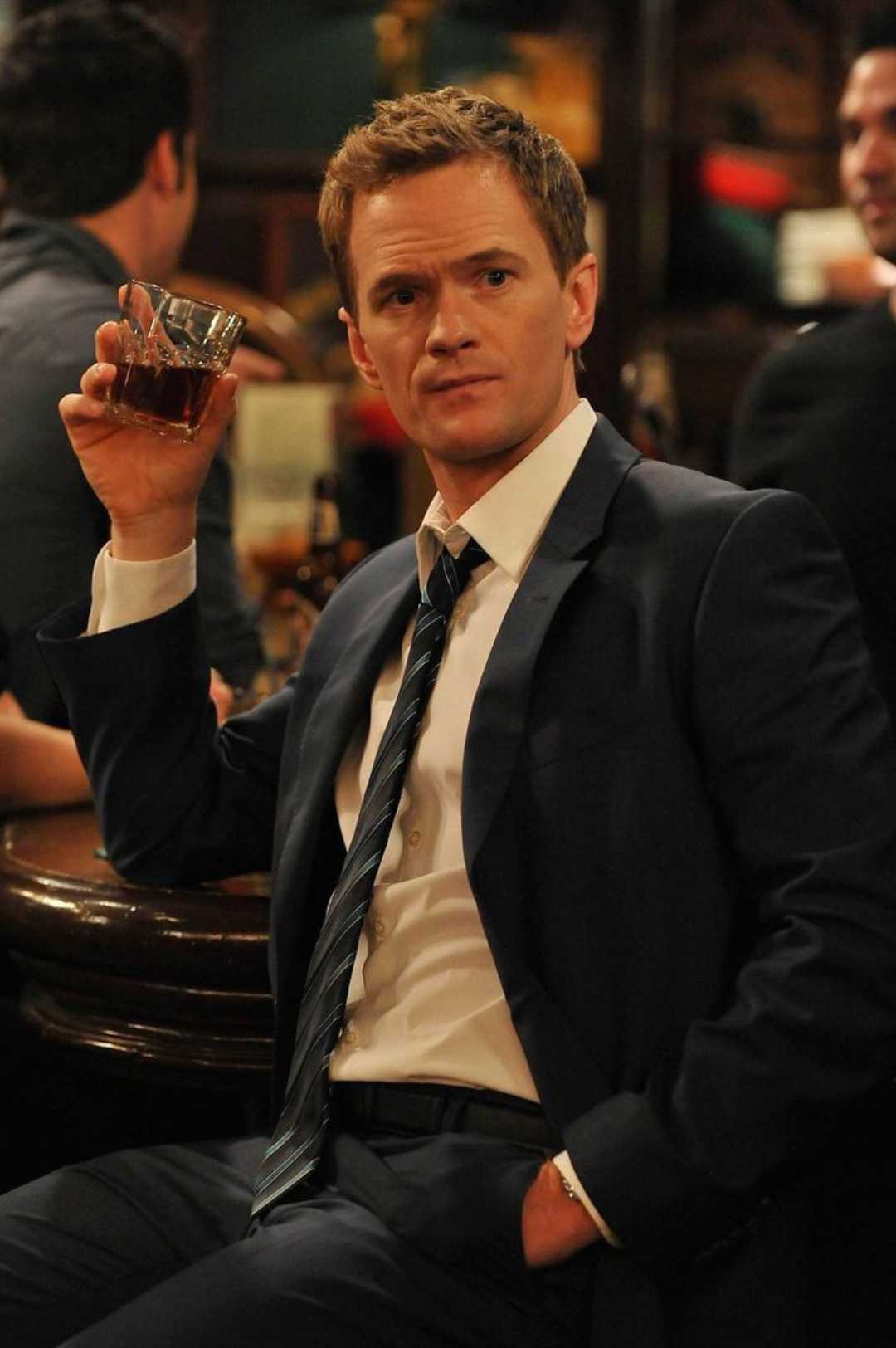 A man in a suit sits at a bar, holding up a glass of whiskey and looking to the side with a thoughtful expression. The background shows other people and a dimly lit bar scene.