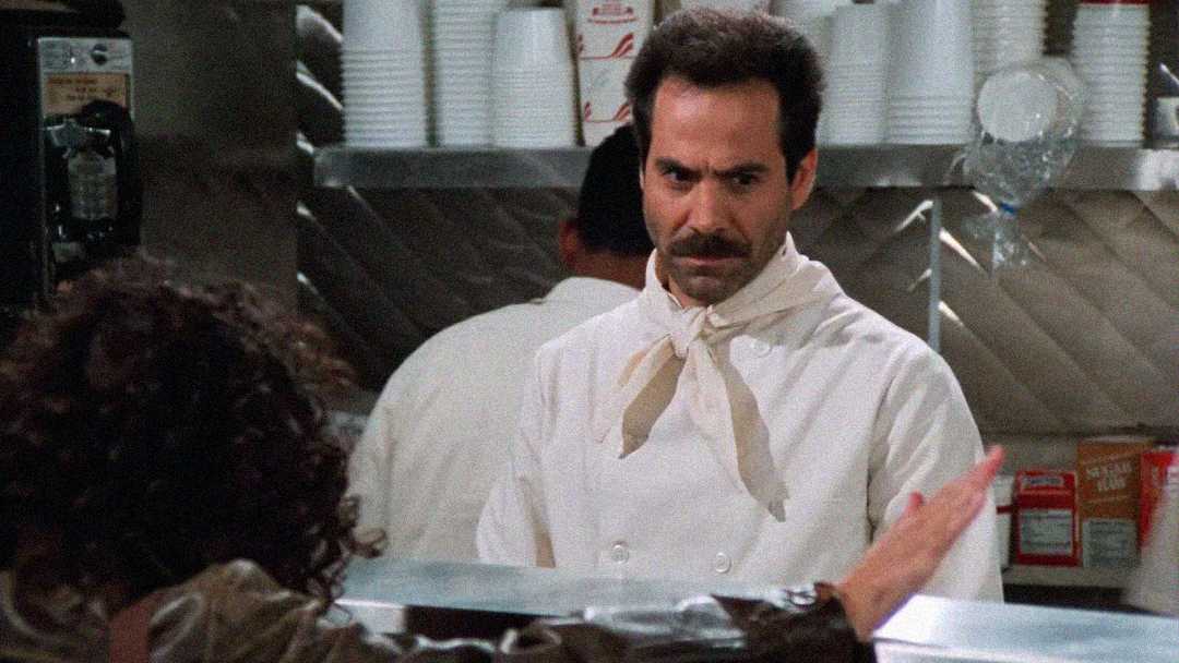 A stern-looking man in a white chef’s coat and neckerchief stands behind a diner counter, glaring at a customer with his arm raised. The kitchen background includes cups and shelves.