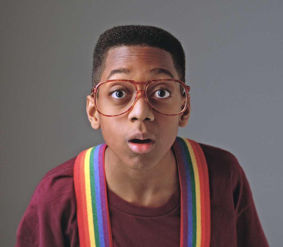 A young boy with a flat-top haircut, wearing large red glasses, a maroon shirt, and rainbow suspenders, looks surprised with his mouth slightly open against a plain gray background.
