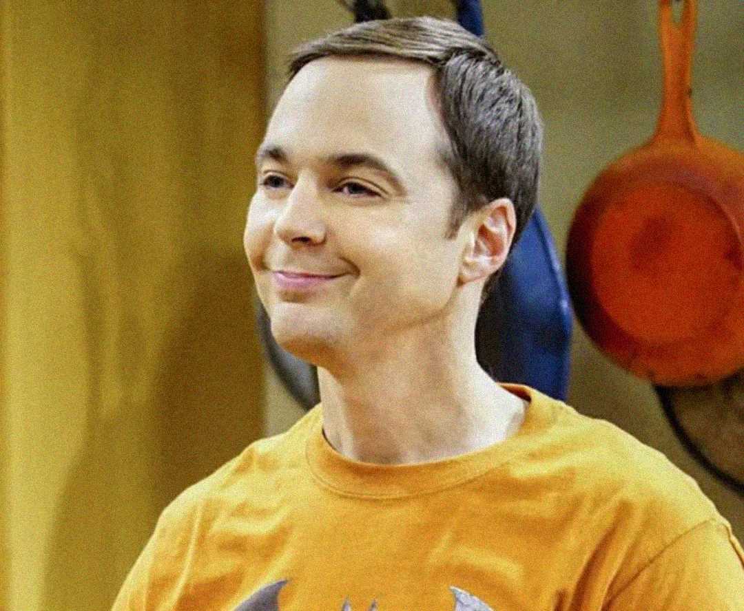 A man with short brown hair is smiling slightly while wearing an orange graphic T-shirt. There are colorful pans hanging on the wall in the background.