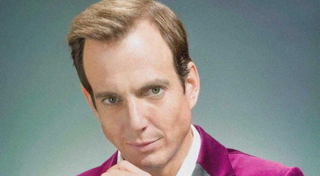 A man with short light brown hair and fair skin gazes thoughtfully at the camera, resting his chin on his hand. He is wearing a bright purple jacket over a light shirt against a neutral background.