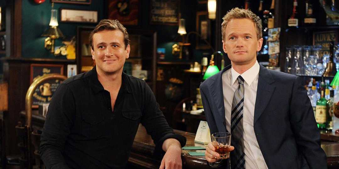 Two men are sitting at a bar. One, dressed casually in a black shirt, is smiling, while the other, in a suit and tie, holds a glass and looks ahead. There are bottles and lamps in the background.