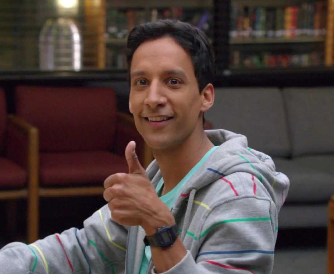 A man with short dark hair wearing a light gray hoodie with colorful stripes smiles and gives a thumbs up while sitting indoors on a chair, with couches and bookshelves in the background.