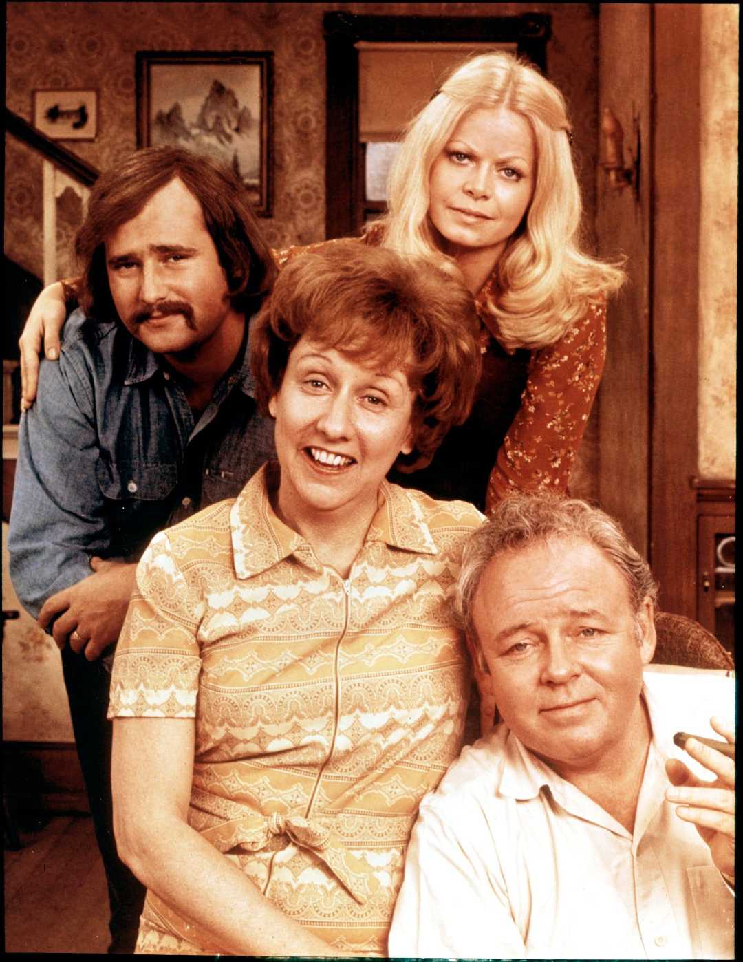 Four adults pose together in a cozy, vintage living room. Two women are in front and back, one smiling with short hair, and one with long blonde hair, while two men sit and stand beside them, all smiling or looking content.