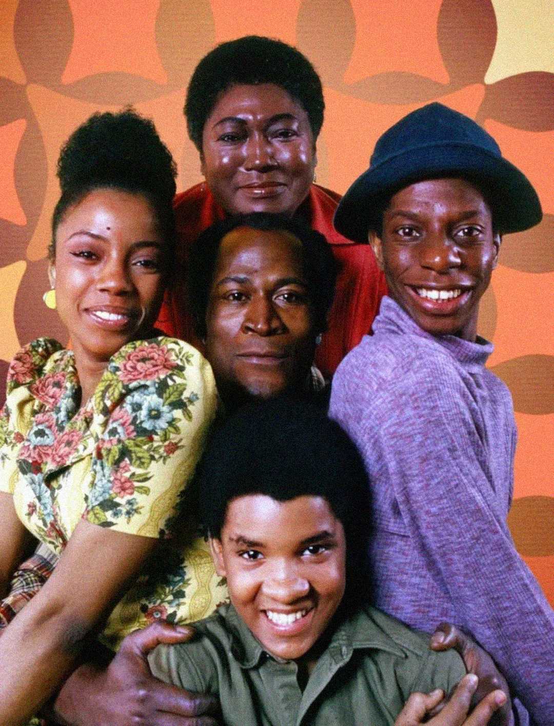 Five people pose together, smiling, in front of an orange patterned background. They are dressed in colorful, vintage clothing reflecting a 1970s style, with close, cheerful expressions.