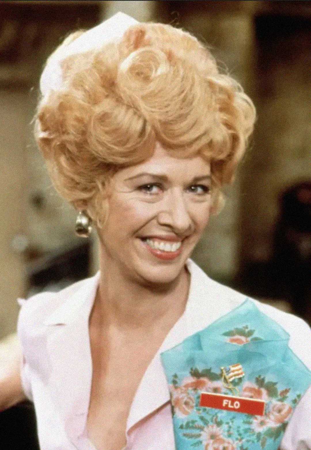 A woman with styled blonde hair, large earrings, and a bright smile wears a light pink uniform. She has a floral apron with a name tag that reads "Flo" and stands in what appears to be a diner or restaurant setting.