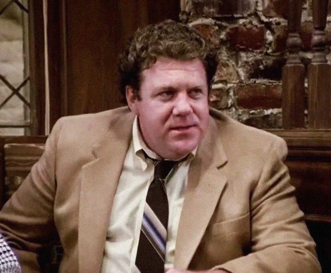 A man with curly hair wearing a tan blazer, white shirt, and striped tie sits indoors in front of a brick wall, looking slightly to his left.