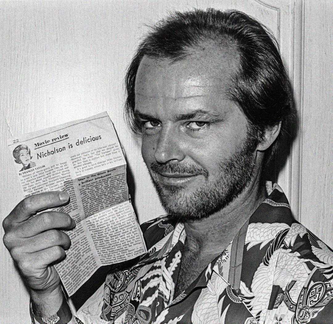 A man with a beard and receding hairline smiles while holding up a newspaper with the visible headline "Nicholson is delicious." He is wearing a patterned shirt and standing in front of a plain wall.