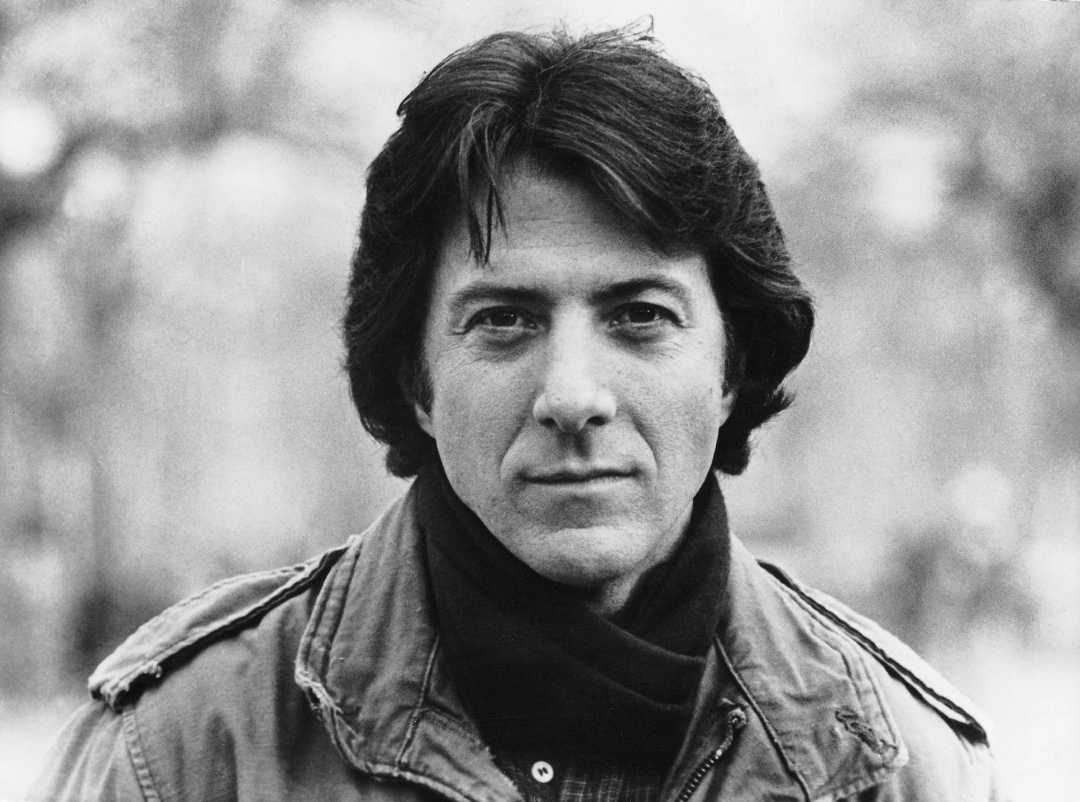 A black and white portrait of a man with dark hair, wearing a jacket and scarf, looking directly at the camera with a neutral expression. The background is blurred with an outdoor setting.