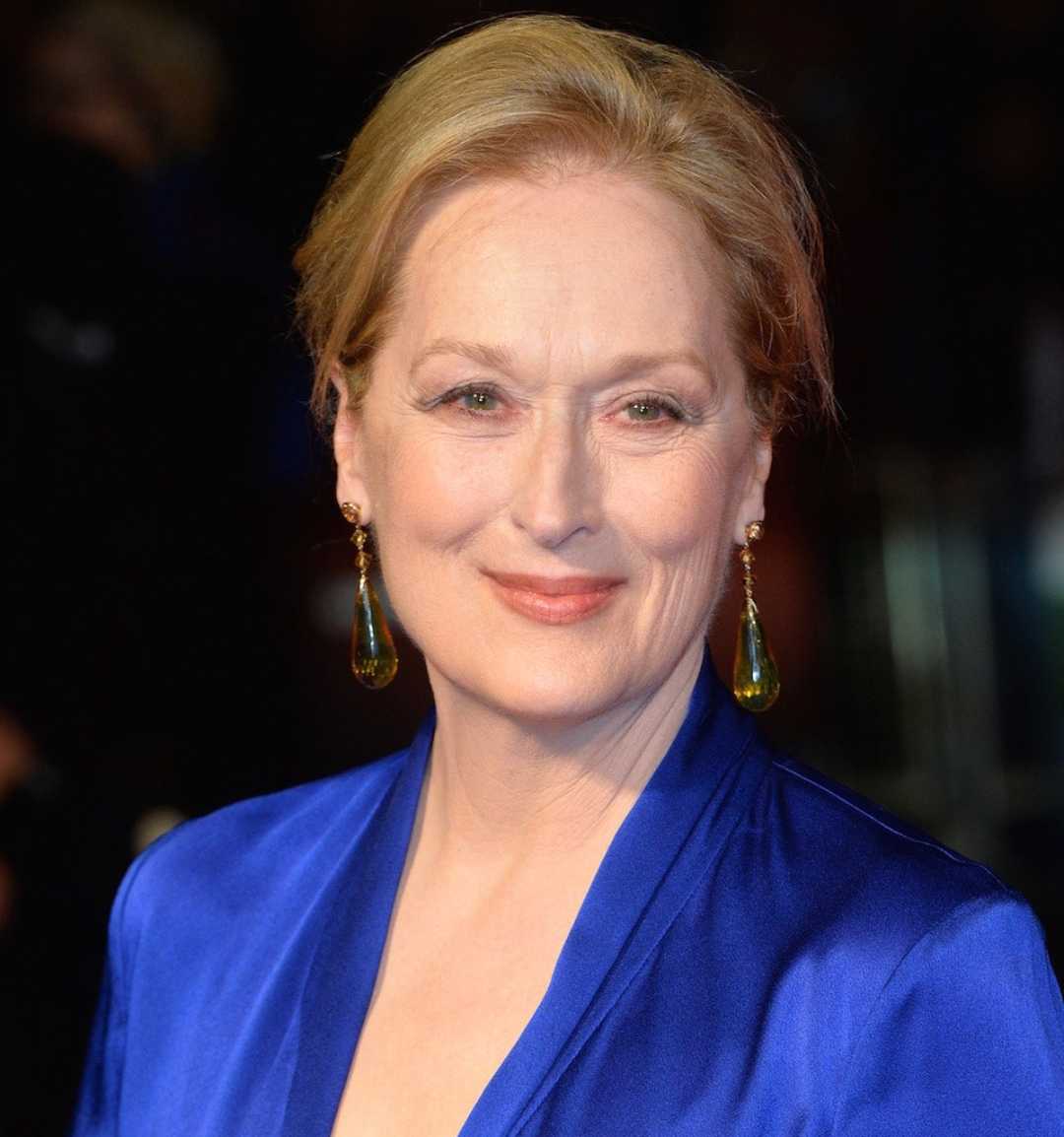 A woman with blonde hair styled back, wearing a royal blue outfit and dangling green earrings, smiles softly at the camera against a dark, blurred background.