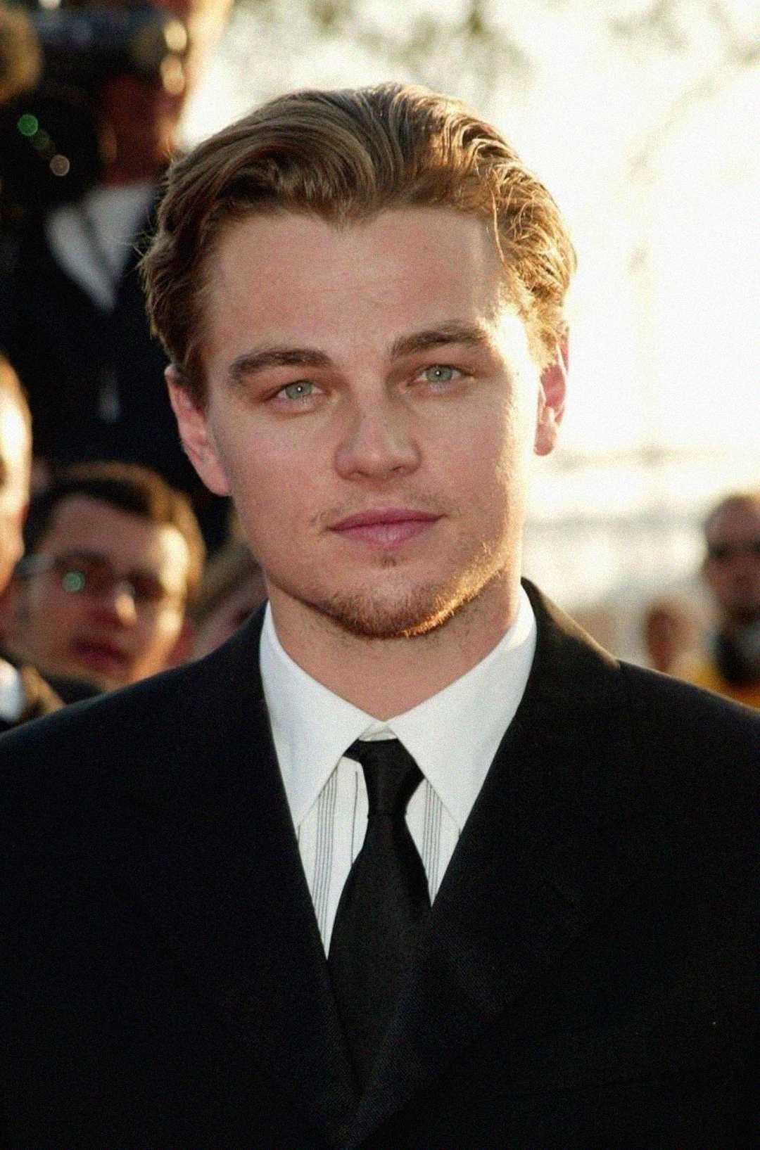 A young man with slicked-back hair wearing a dark suit, white shirt, and black tie is standing outside, looking at the camera. The background is blurred with people and sunlight visible.