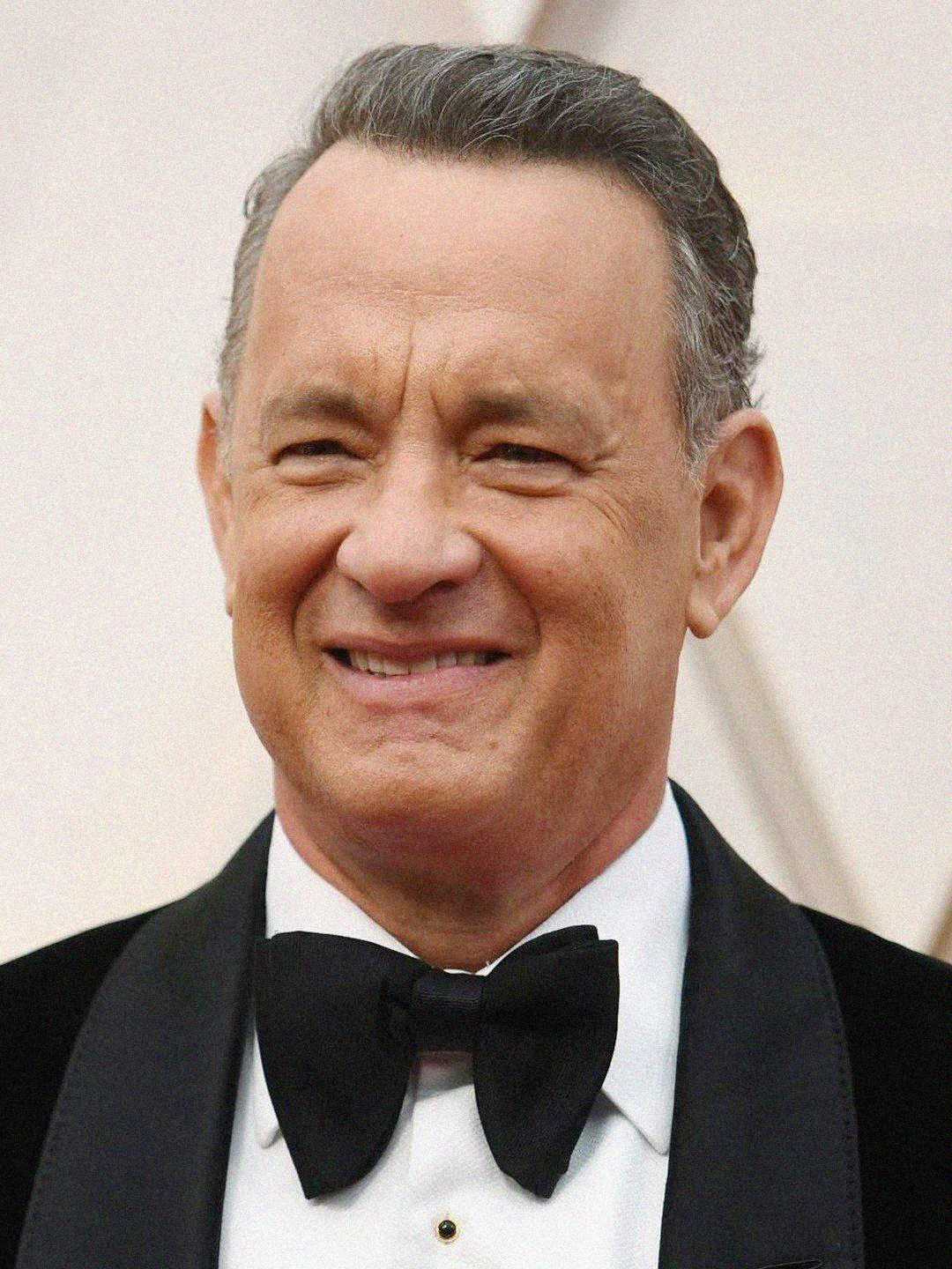 A man with short gray hair is wearing a black tuxedo and bow tie, smiling at the camera against a blurred, light-colored background.