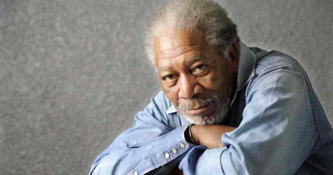 An older man with gray hair and a beard, wearing a light blue denim shirt, rests his arms on a surface and looks thoughtfully at the camera against a plain, textured gray background.