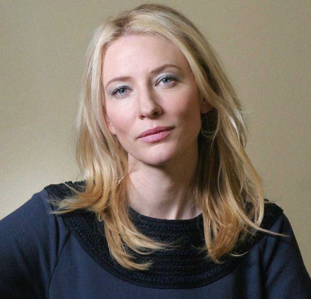A woman with long, blonde hair and fair skin is wearing a dark top. She has a neutral expression and is sitting in front of a plain, light-colored background.