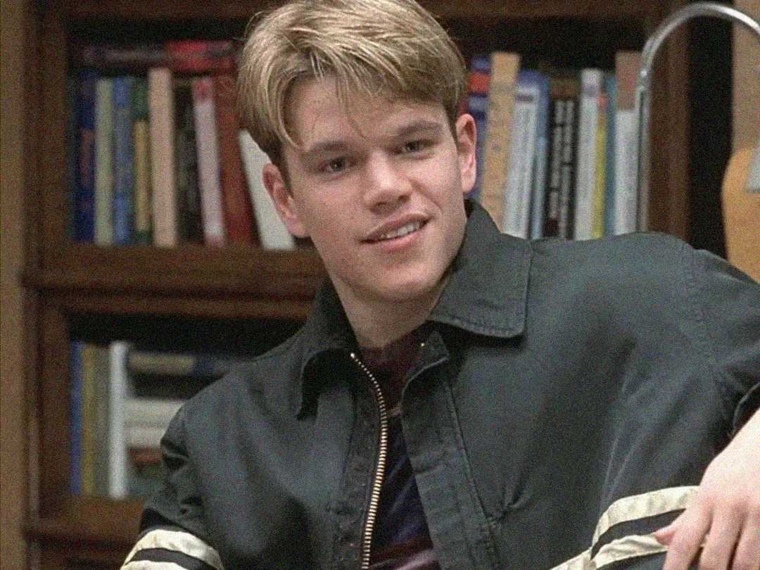 A young man with light brown hair, wearing a dark jacket with light stripes on the sleeves, sits in front of a bookshelf and smiles slightly.