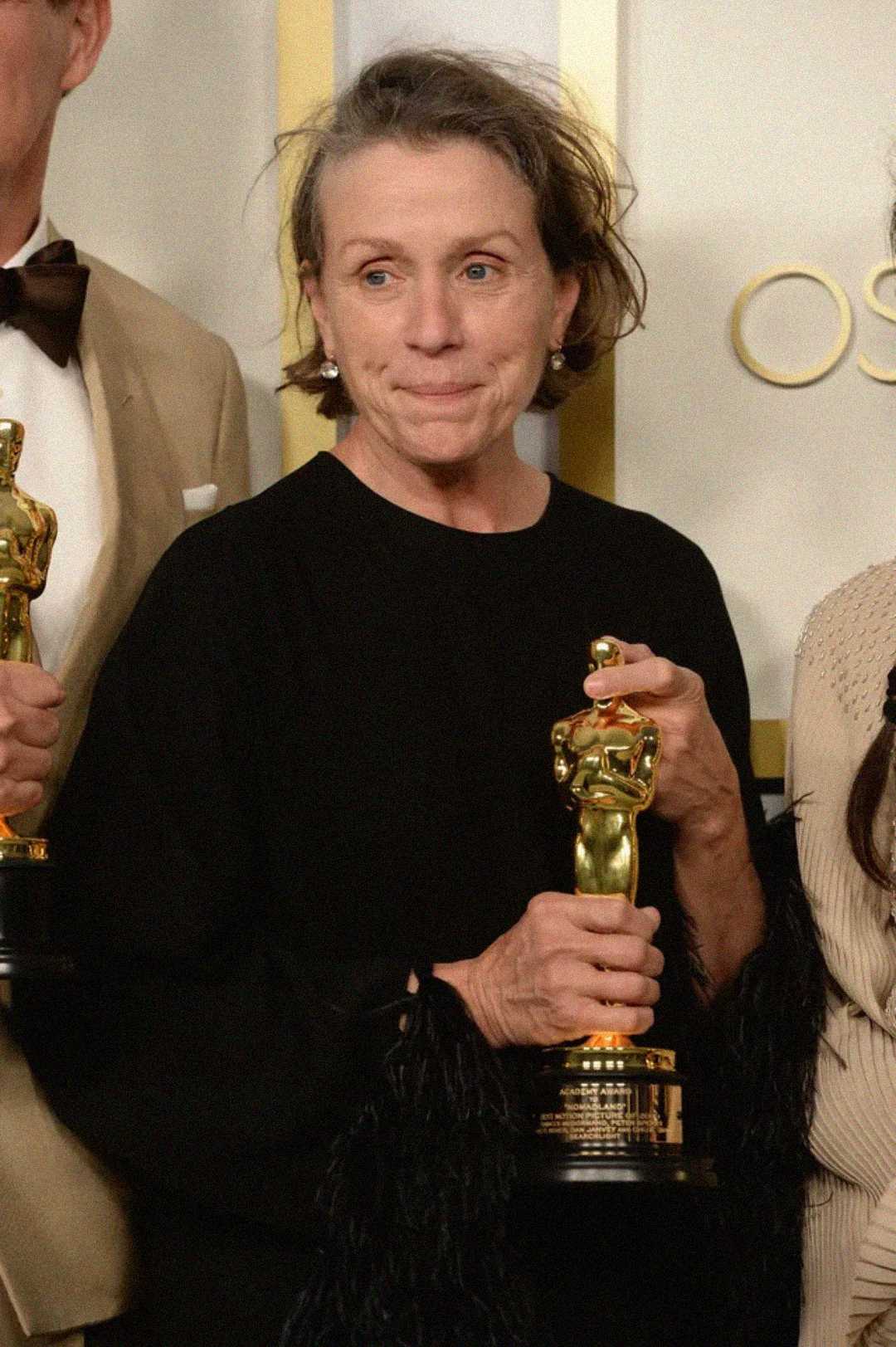 A woman in a black outfit holds an Oscar trophy, standing in front of a neutral backdrop with gold lettering. She has short, tousled hair and a neutral expression. Other people holding Oscars are partially visible beside her.