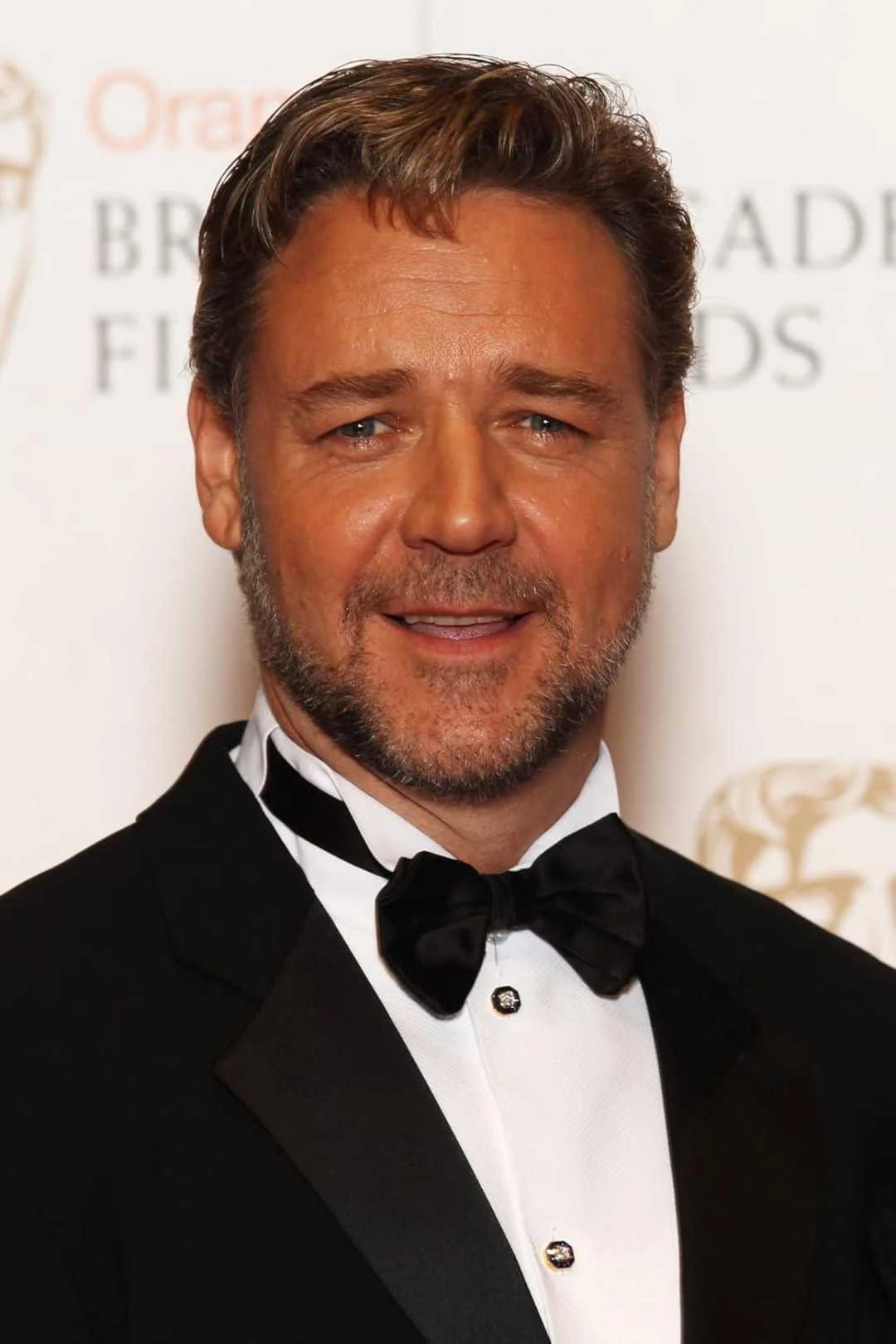 A man with short brown hair and a trimmed beard is wearing a black tuxedo with a black bow tie, standing in front of a white background with partially visible text and logos.