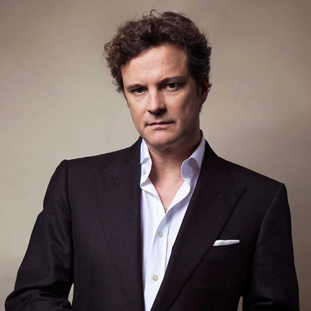 A man with short curly brown hair, wearing a black suit jacket and white dress shirt, stands against a plain beige background, looking directly at the camera with a neutral expression.