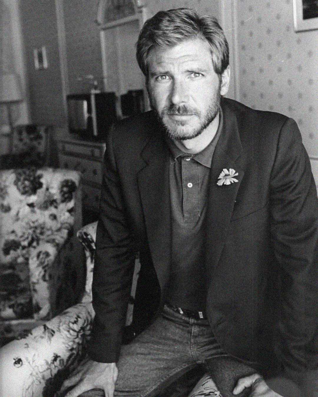 Black and white photo of a man with a beard and short hair, wearing a blazer with a flower pin and a polo shirt, sitting on the arm of a floral armchair in a vintage-style room.