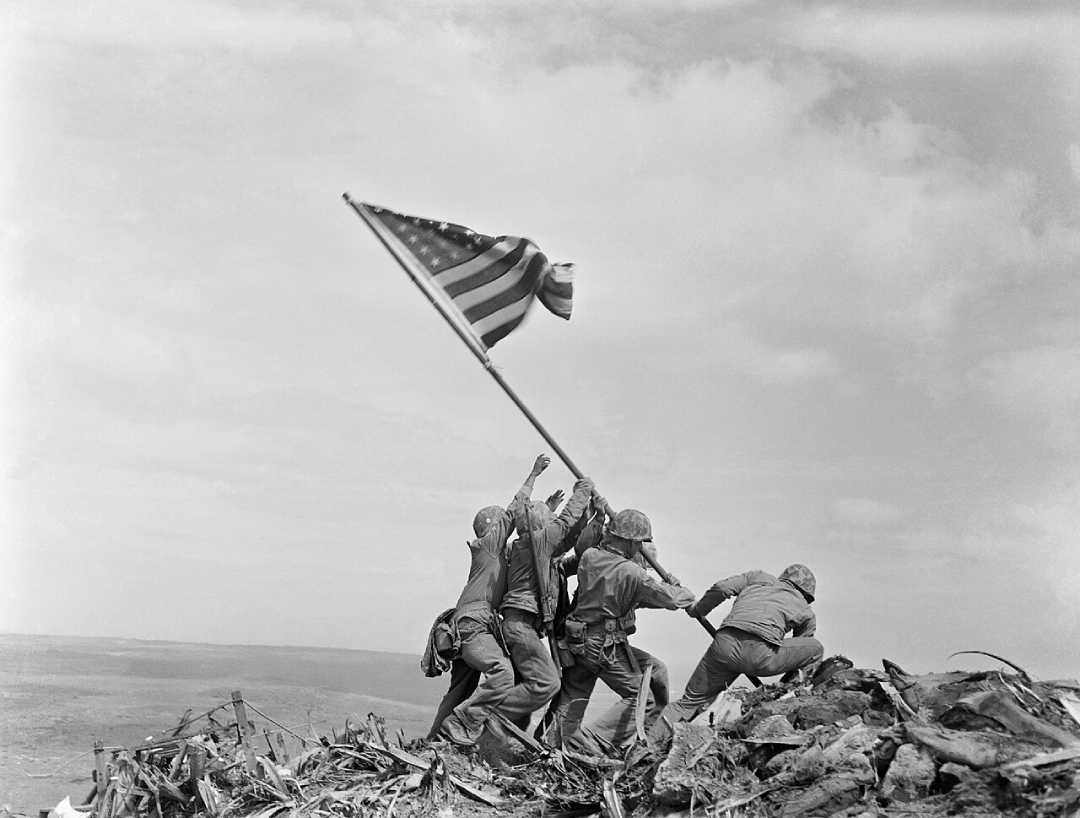 Five soldiers raise an American flag together atop a rocky hill during daylight, with a cloudy sky in the background. The scene conveys teamwork, struggle, and victory.