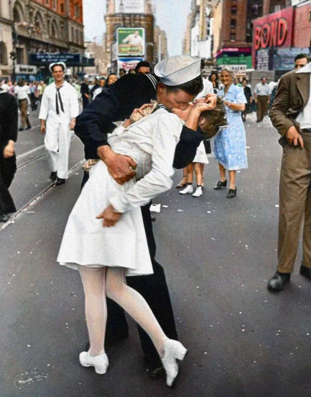 A sailor passionately kisses a woman in a white dress on a busy city street, surrounded by people celebrating. The scene is lively, with vintage buildings and a "BOND" sign visible in the background.