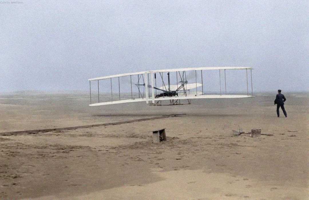 A vintage biplane with a wooden frame flies low over a sandy, barren landscape as a person stands nearby, watching its flight. The scene appears historical and pioneering.