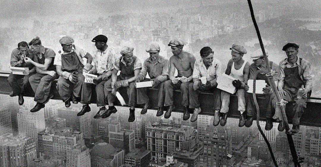 Eleven construction workers eat lunch and chat, sitting on a steel beam high above New York City, with skyscrapers visible far below. The image is in black and white.