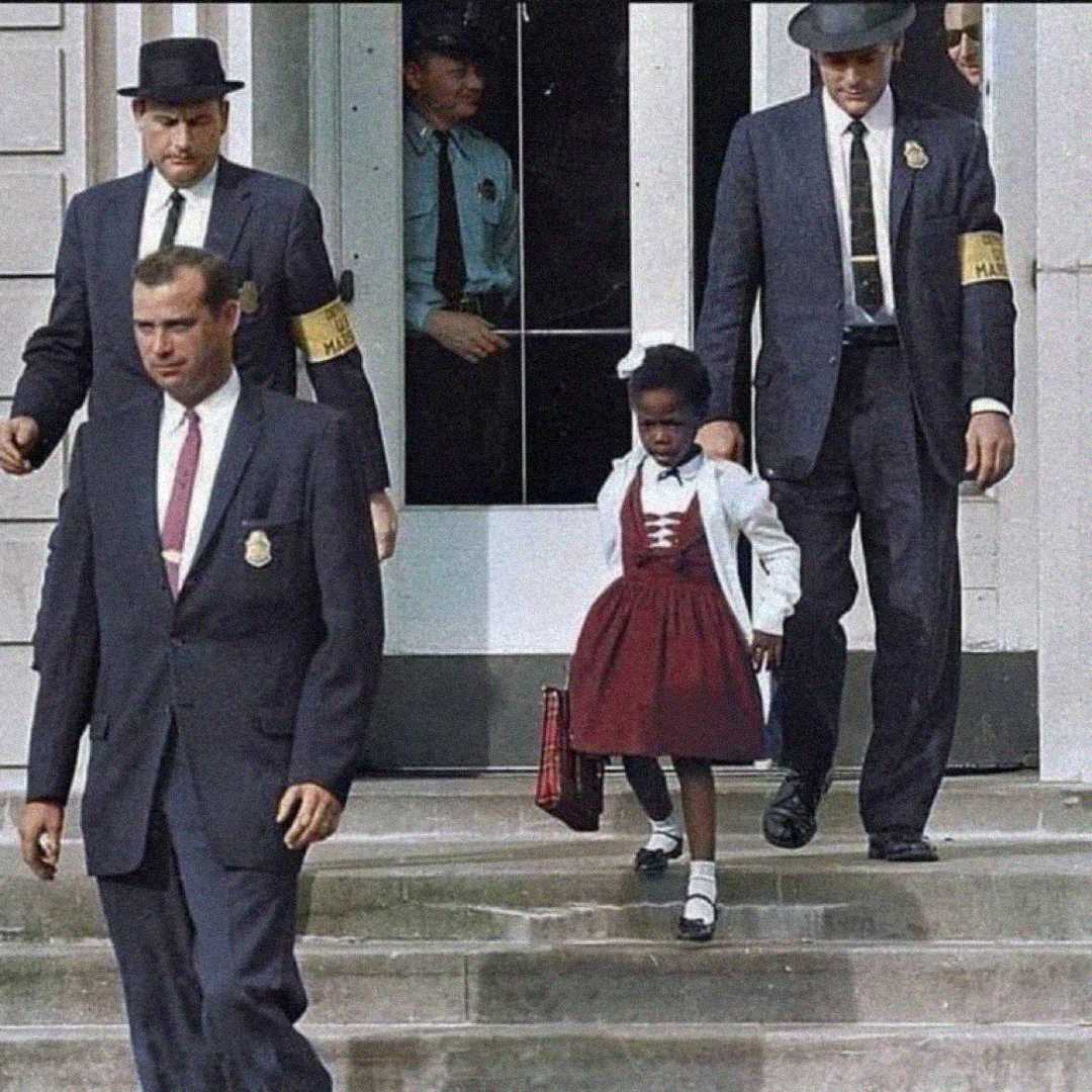 A young Black girl in a red dress carries a school bag while being escorted by four suited federal marshals outside a building, with onlookers visible in the doorway behind them.