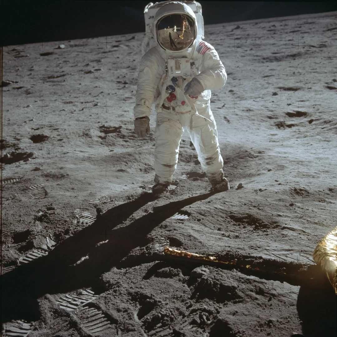 An astronaut in a white spacesuit stands on the gray, dusty surface of the Moon, with footprints and equipment visible, and their reflection appears in their helmet visor.