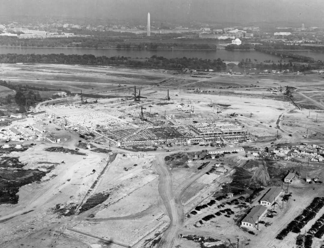 Aerial view of a large construction site near a river, with the Washington Monument and the Lincoln Memorial visible in the background across the water in Washington, D.C.