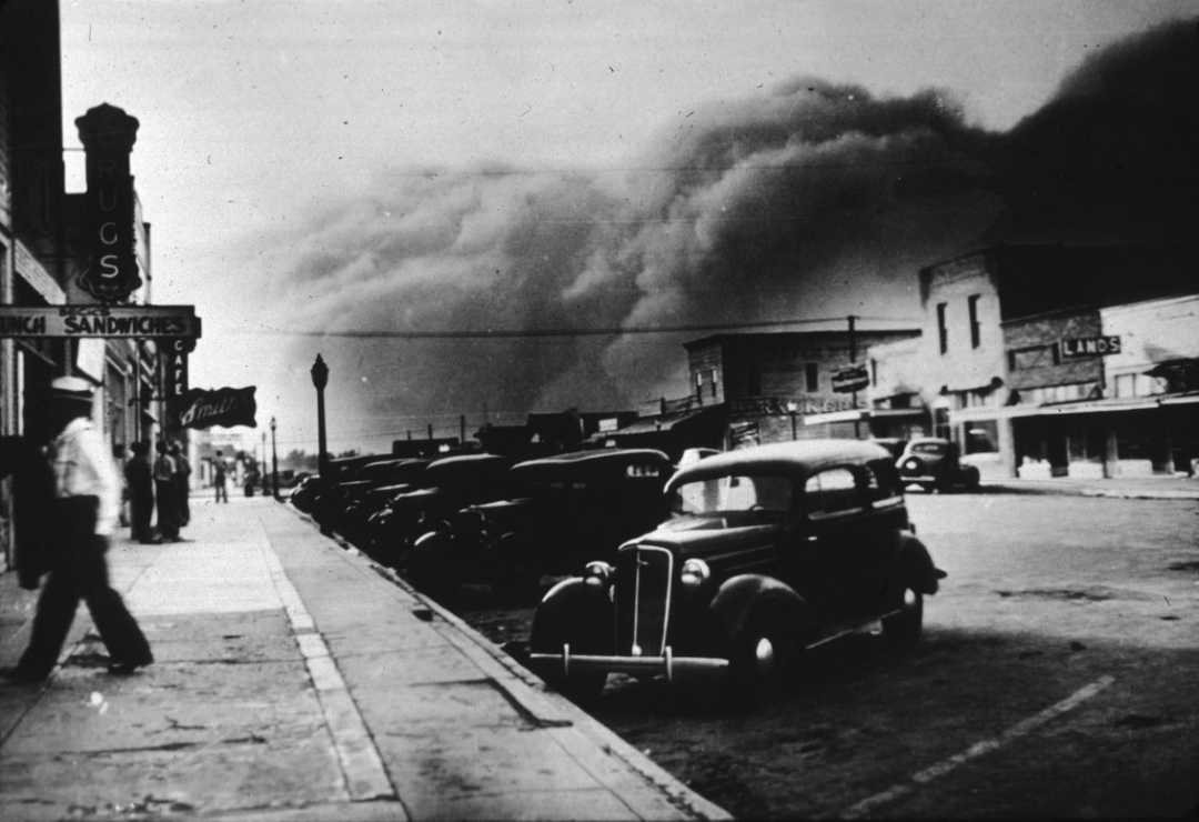 A black-and-white photo shows a main street lined with parked 1930s cars as a large dust storm approaches, filling the sky with dark clouds. A few people walk along the sidewalk beside storefronts.