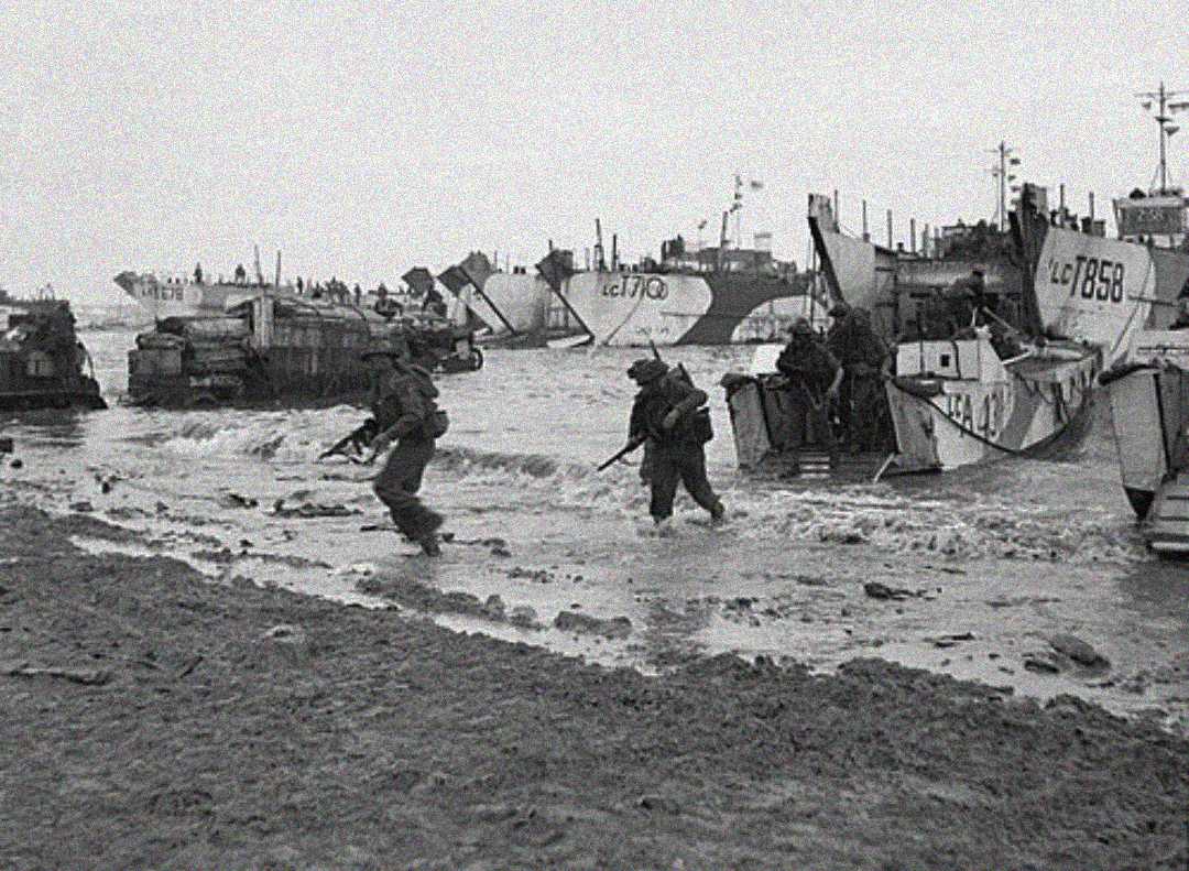 Black and white photo of soldiers with rifles landing on a beach from military boats. Several landing crafts are in shallow water, and soldiers are wading ashore amid scattered equipment. The scene appears tense and chaotic.
