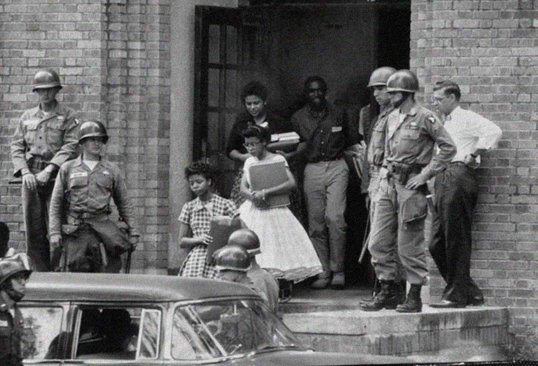 A group of Black students exits a brick building, holding books and notebooks, escorted by armed soldiers in helmets. Onlookers stand nearby, and a car is parked in the foreground. The scene appears tense and historic.