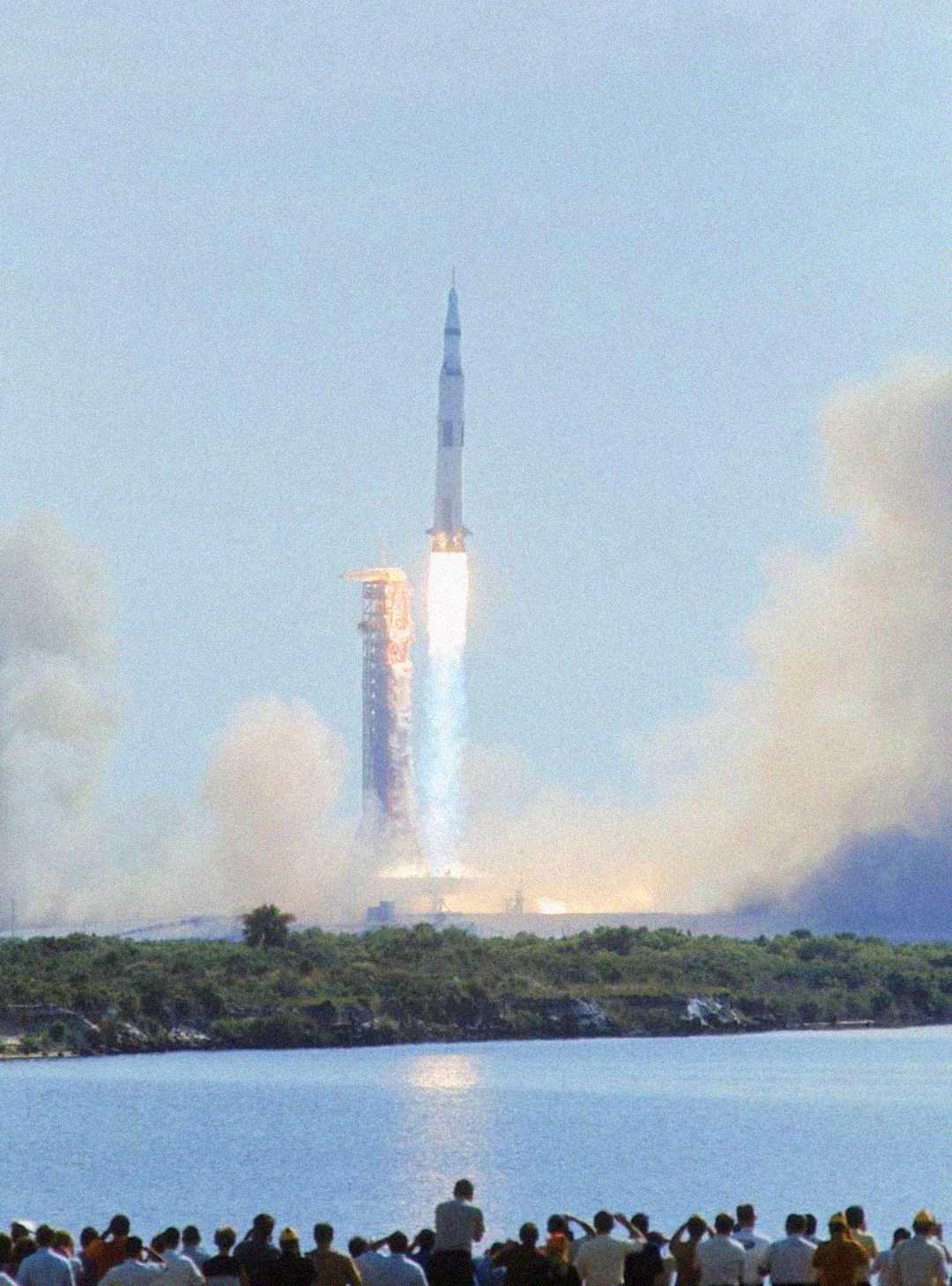A large crowd watches from across the water as a rocket launches, sending bright flames and smoke billowing into the sky against a pale blue background.