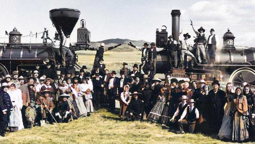 Large group of men and women in 19th-century clothing pose in front of two old steam locomotives on a grassy field, with mountains visible in the background on a partly cloudy day.