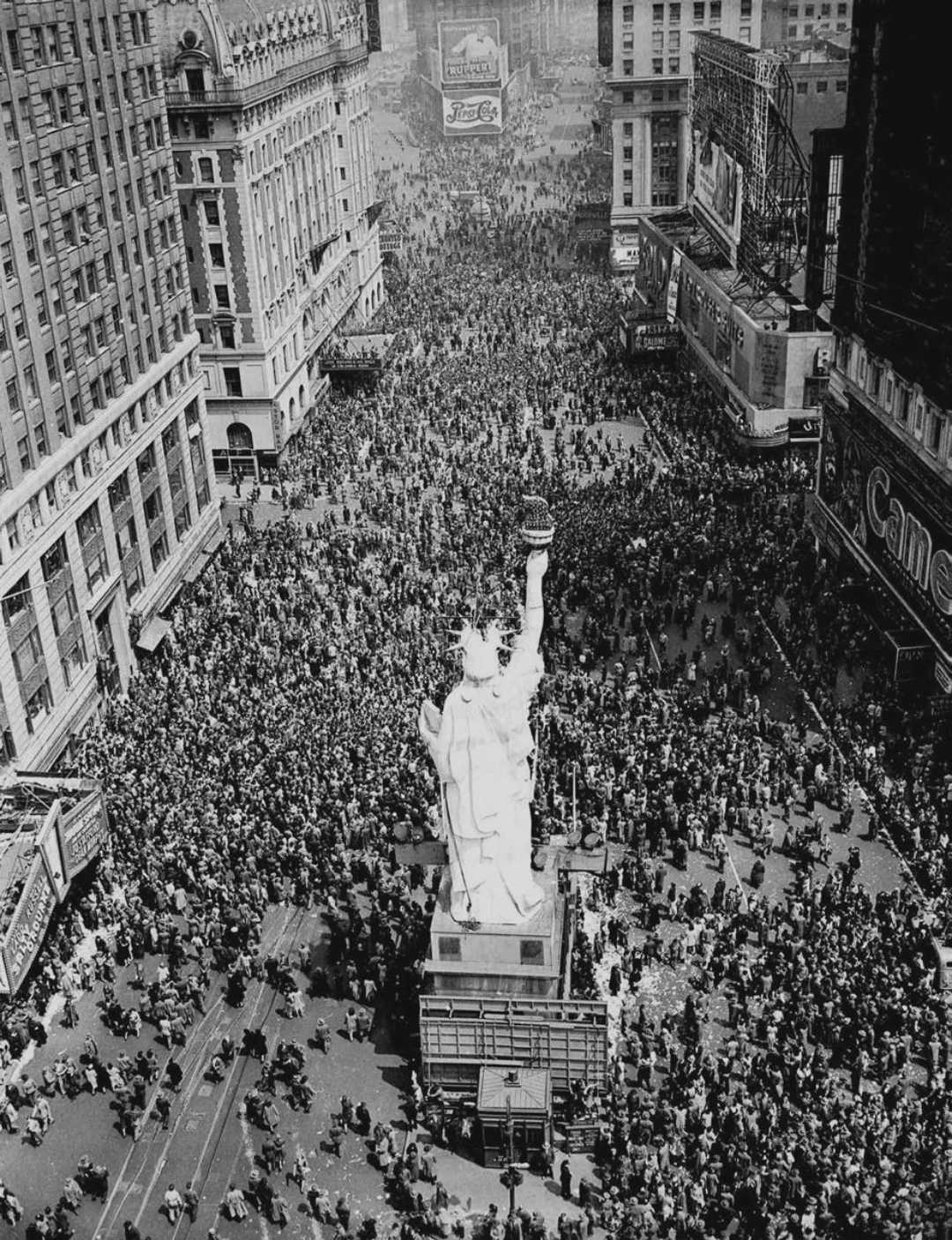 A large crowd fills Times Square in New York City for V-E Day celebrations in 1945. A replica of the Statue of Liberty stands in the center, surrounded by people and tall buildings.