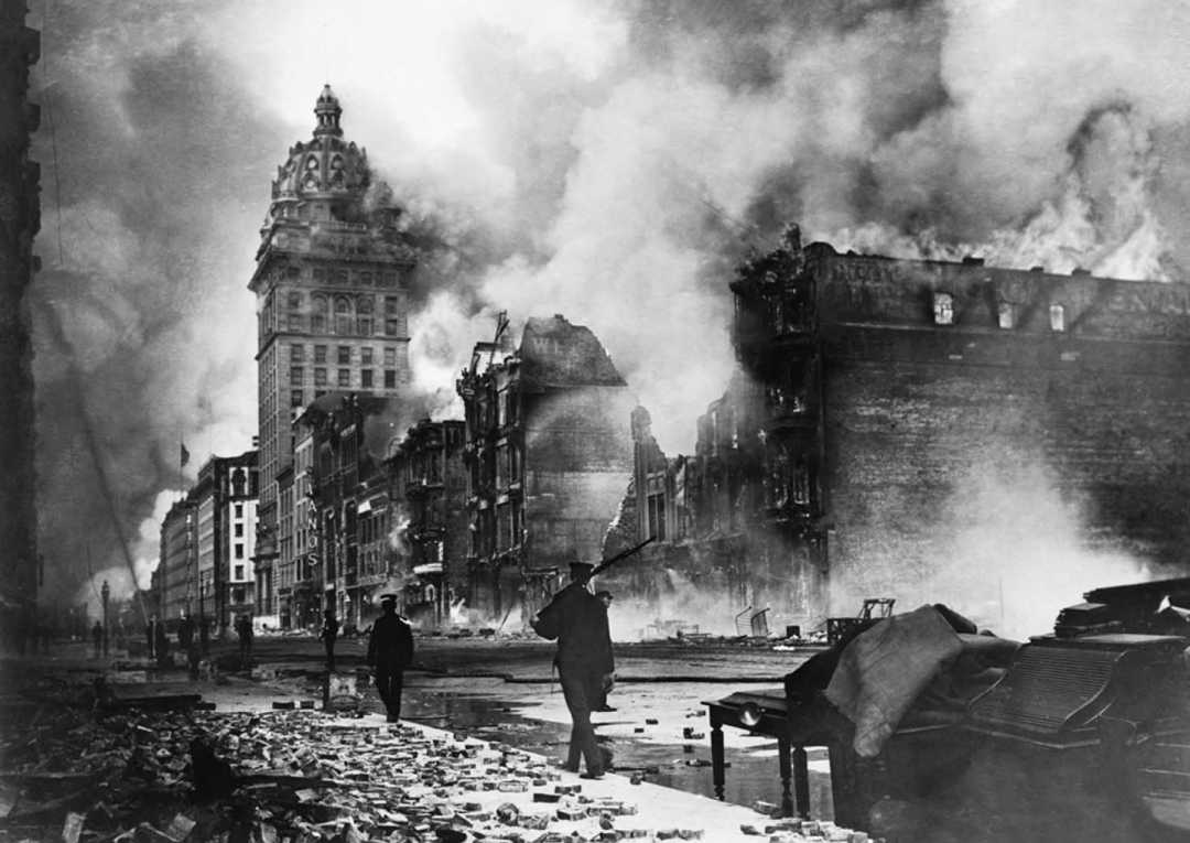 Smoke and flames rise from destroyed buildings along a city street, with debris scattered everywhere. A few people walk amid the devastation. A tall, ornate building stands in the background, mostly intact.