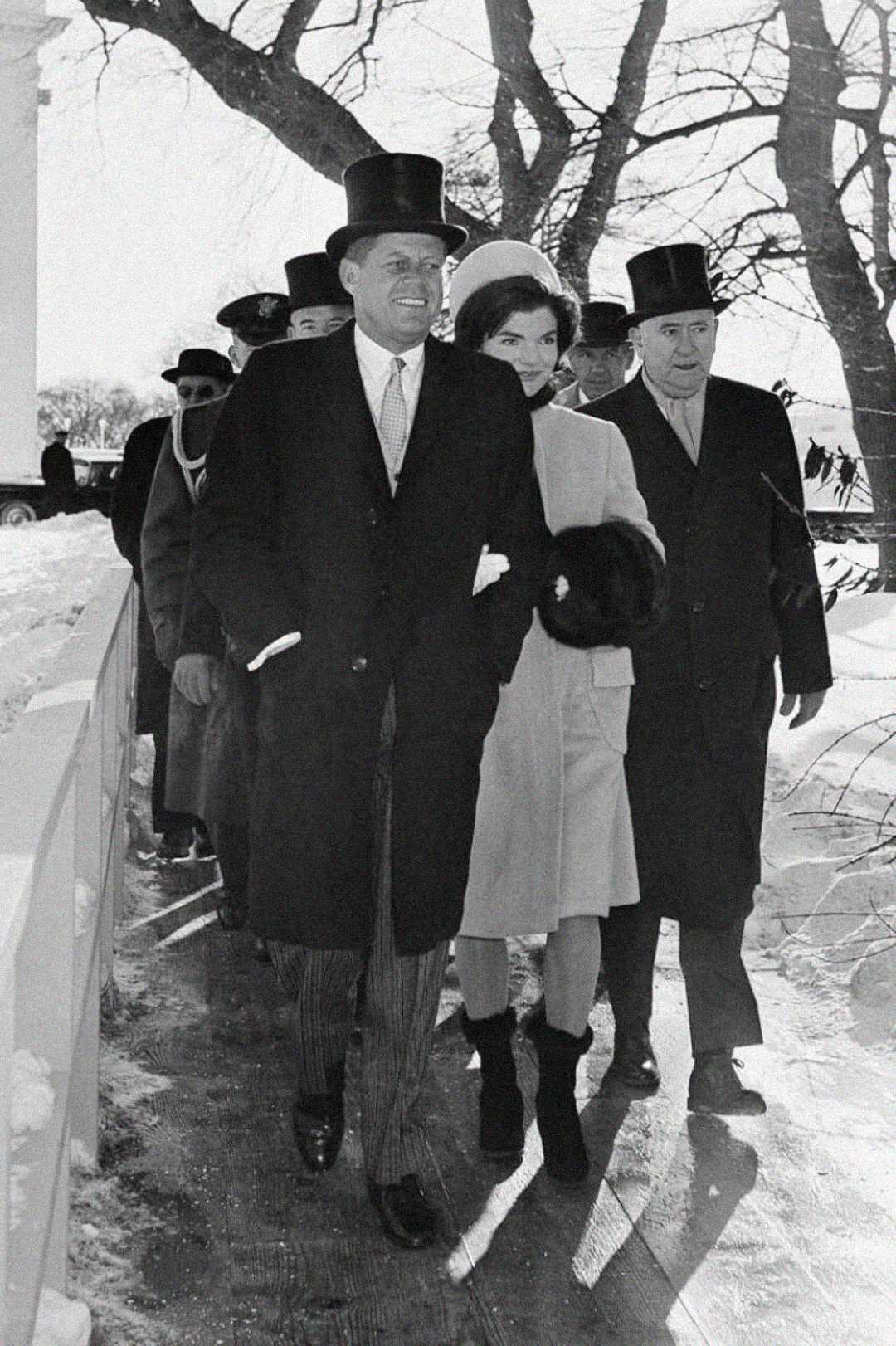 A group of well-dressed people in winter coats and top hats walk outside on a snowy day, with bare trees in the background. The woman in front wears a light coat and carries a fur muff.