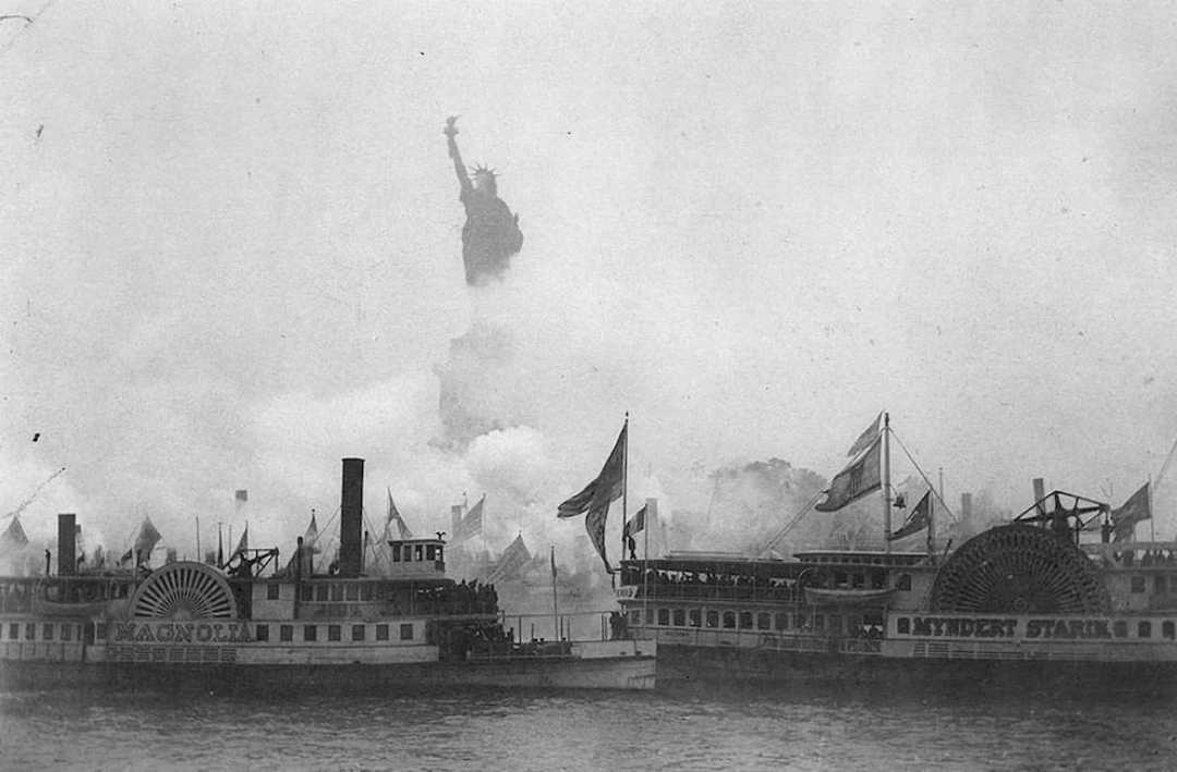 Historic black and white photo of two steamboats on water with the Statue of Liberty visible in the background, partially shrouded in smoke or fog. Crowds and flags are seen on the boats.