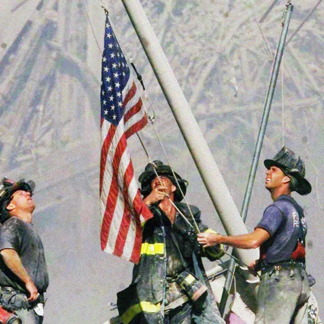 Three firefighters in helmets and gear raise an American flag on a pole amid debris and smoke, symbolizing resilience and unity in the aftermath of a disaster.