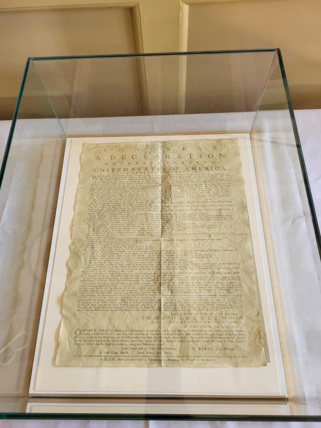 A historical document, the Declaration of Independence, is displayed under a glass case on a white table, mounted on a white mat for preservation and viewing.
