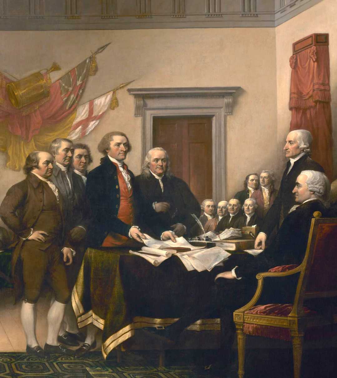 A group of early American leaders in 18th-century attire stand around a table with documents, some seated and some standing, in a formal room with flags and a large red curtain in the background.