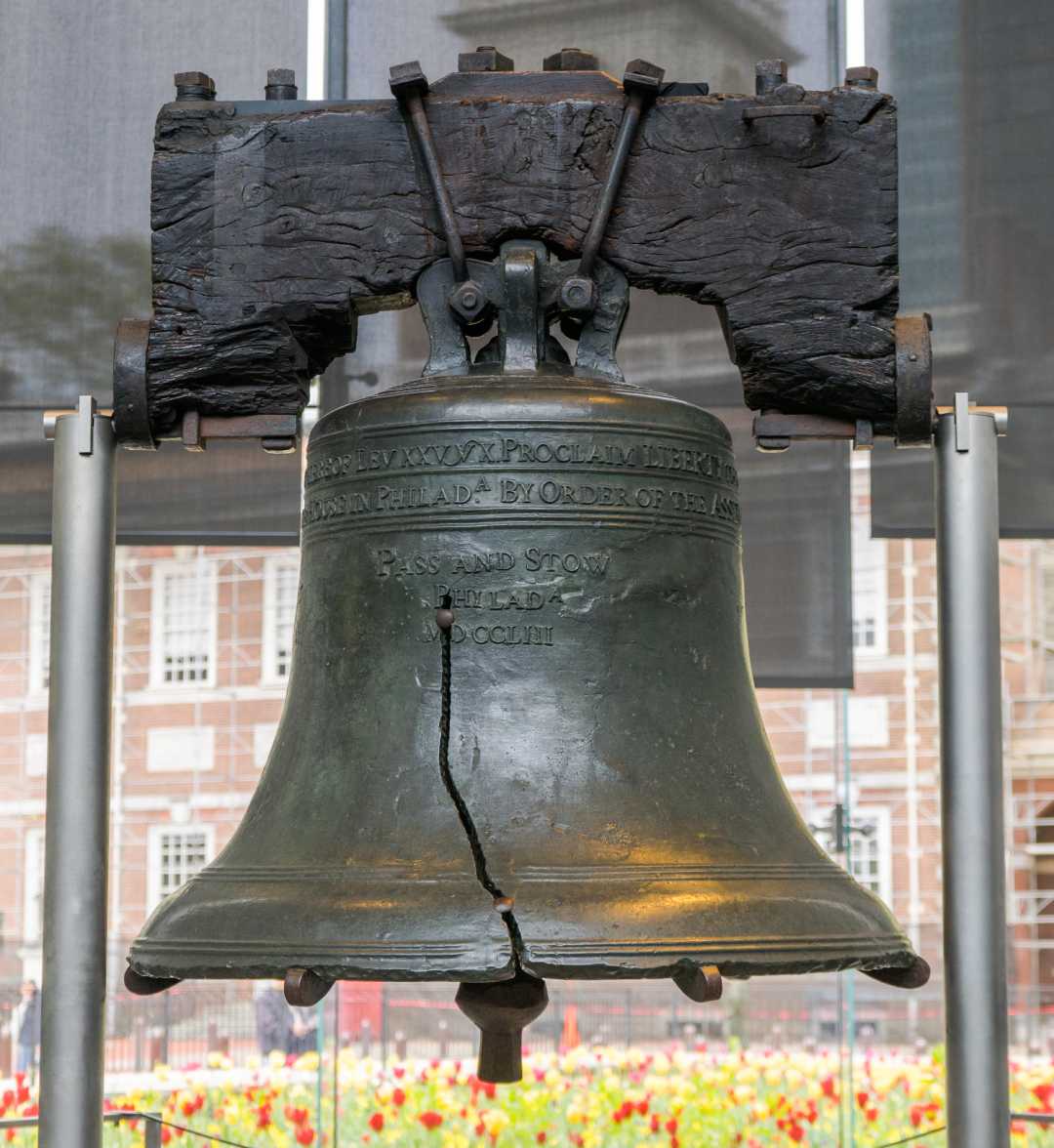 The Liberty Bell, an old bronze bell with a large crack, is displayed indoors on a metal stand with Independence Hall visible in the background.