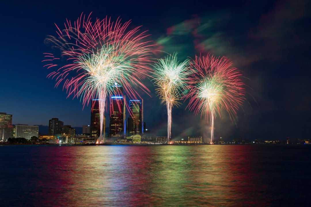 Colorful fireworks burst in the night sky over a city skyline, reflecting vibrant reds, greens, and yellows on the calm water below, with tall buildings illuminated in the background.