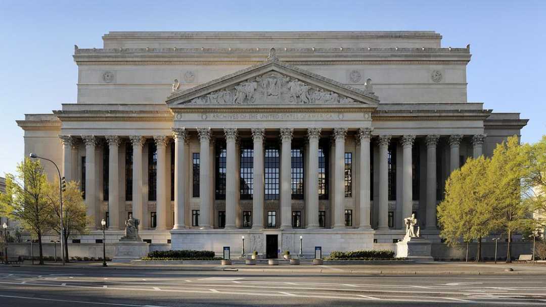 A neoclassical building with tall columns, large stairs, and statues at the entrance, set against a clear blue sky with trees on either side.