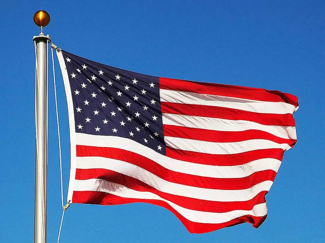 The United States flag waving in the wind on a flagpole, featuring 50 white stars on a blue field and 13 red and white stripes, set against a clear blue sky.